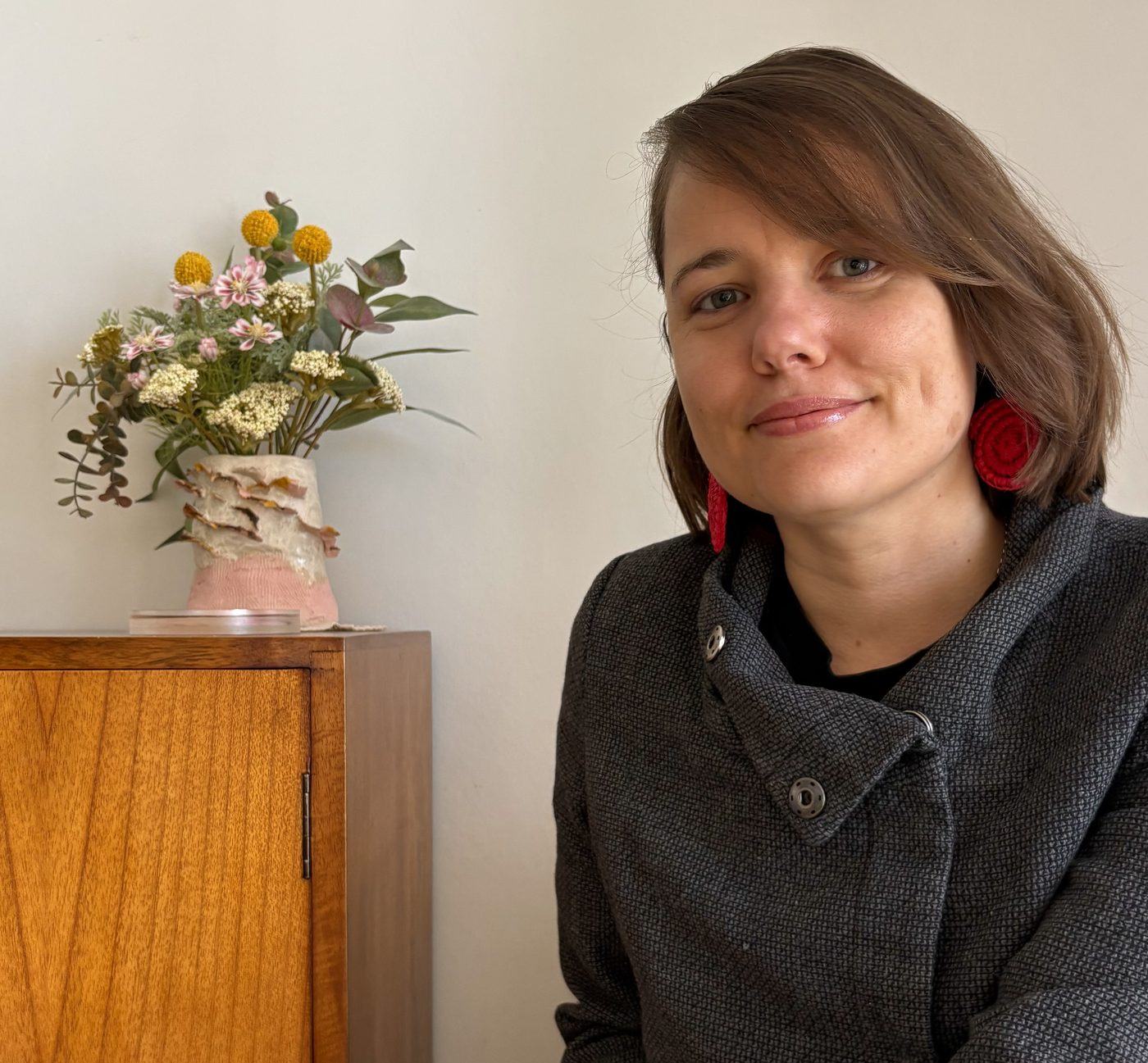 Miranda seated beside a vintage cabinet with a vase of dried flowers